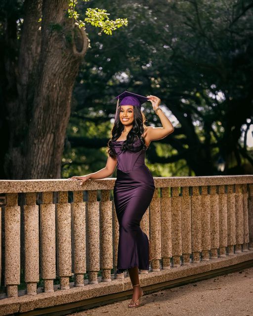 a woman in a graduation gown leaning on a fence with her hands behind her head