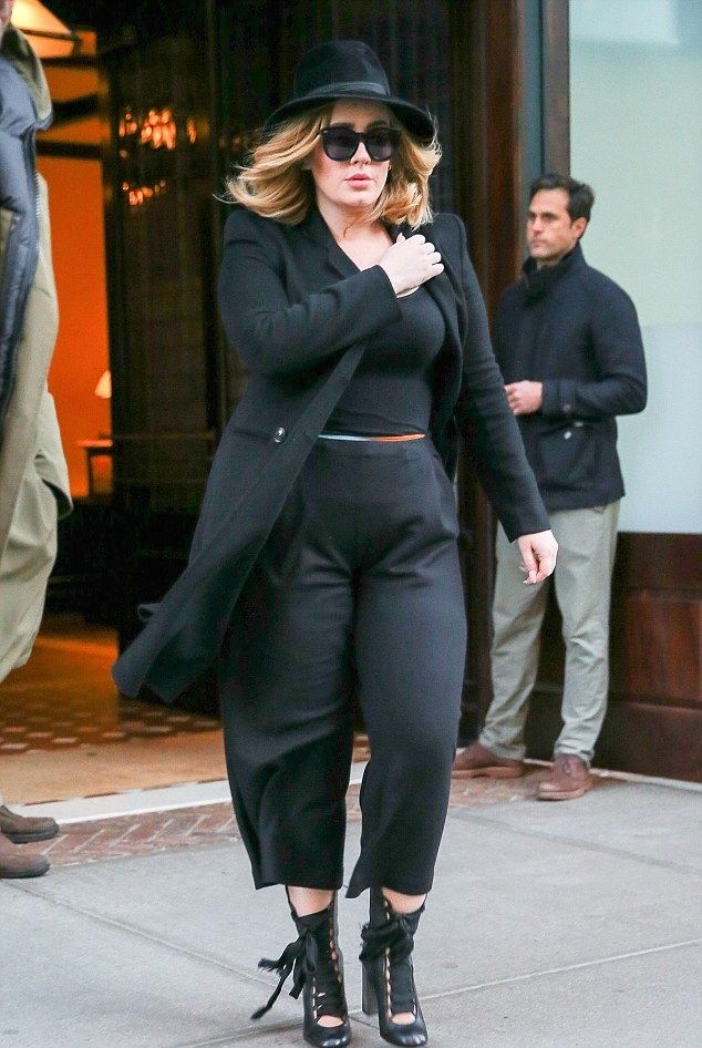 a woman in black outfit and hat walking down the street with her hand on her hip