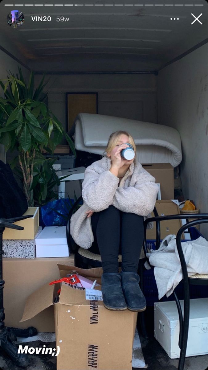 a woman sitting on top of a cardboard box drinking from a cup in her living room