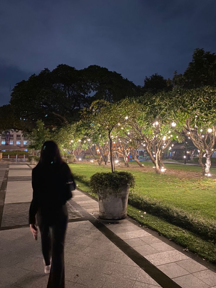 a woman walking down a sidewalk at night with lights on trees and bushes in the background