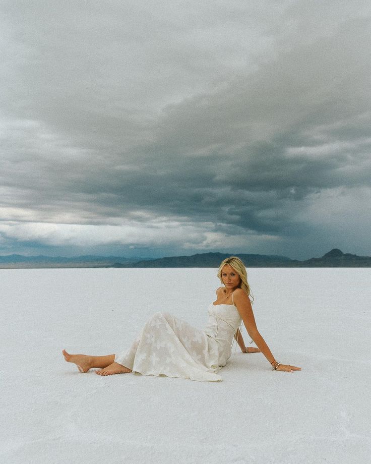 a woman sitting in the middle of an empty field under a cloudy sky with her legs crossed