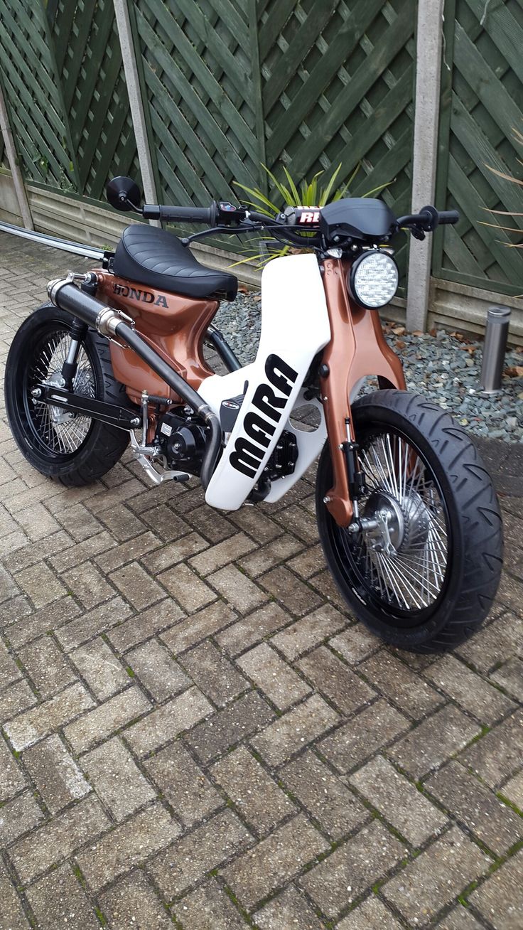 an orange and white moped parked on top of a brick road next to a fence