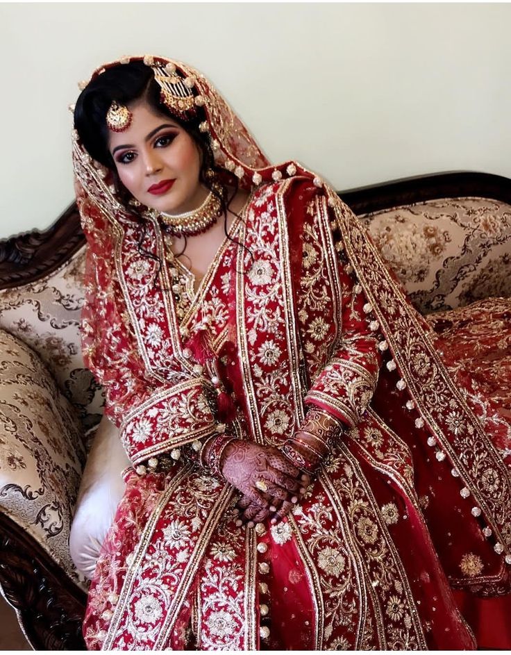 a woman in a red and gold bridal gown sitting on a couch with her hands clasped