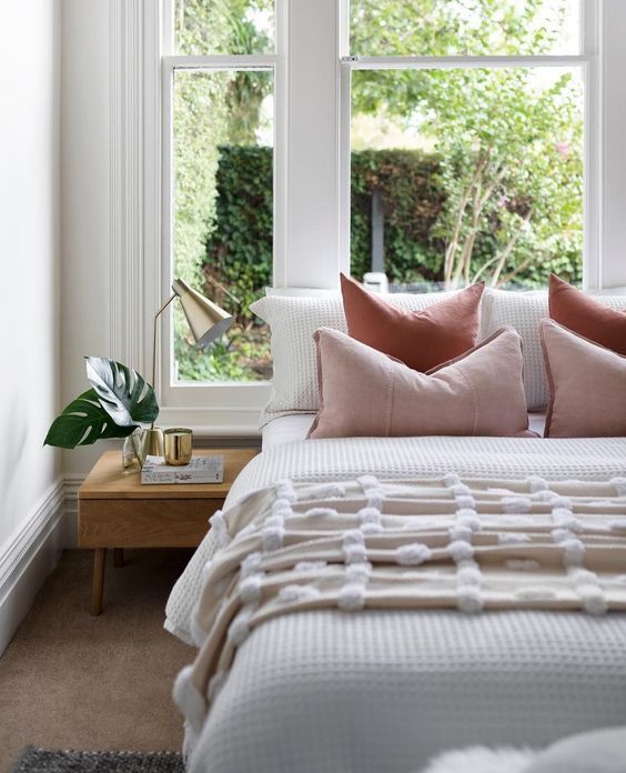 a bed with pink and white pillows in front of a window next to a night stand