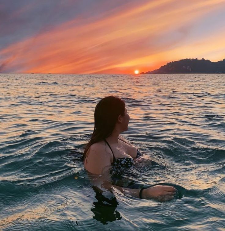 a woman sitting in the water with her arm out and looking into the distance at sunset