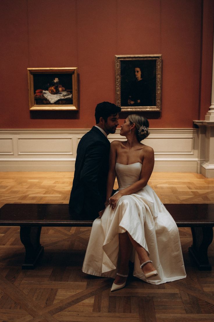 a bride and groom sitting on a bench in an art gallery with paintings behind them