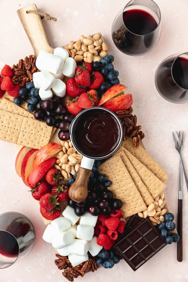 a platter with fruit, crackers, nuts and chocolate on it next to two glasses of wine