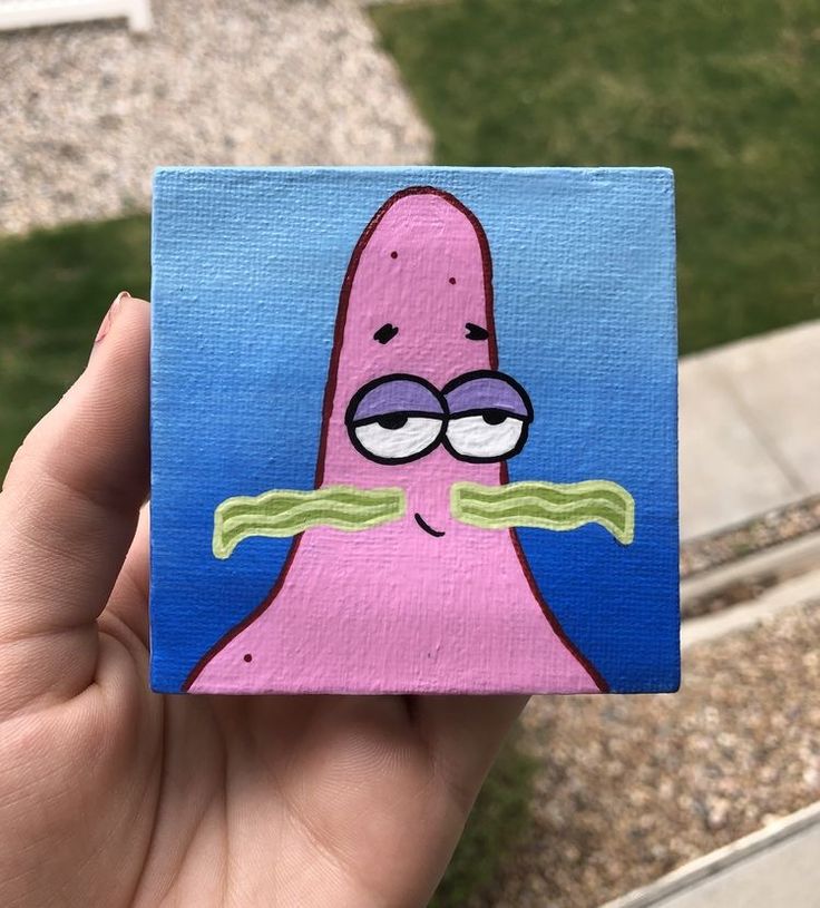 a hand holding up a small square painting of a pink cartoon character with glasses on it's face