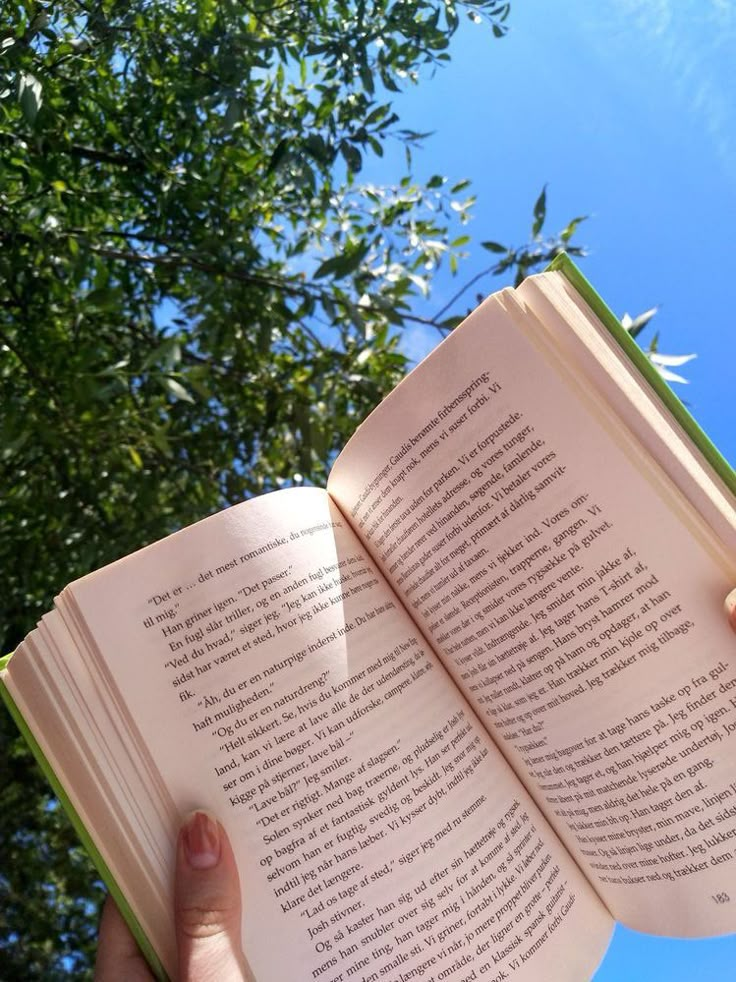 a person holding an open book in their hand with trees in the background and blue sky