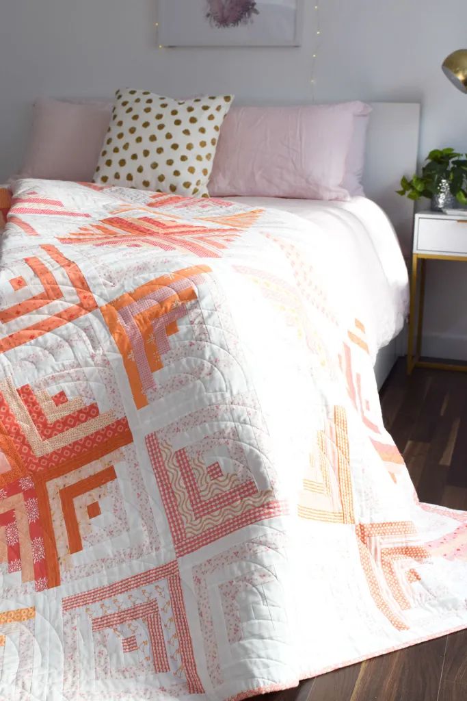 a white bed topped with a pink and orange quilt next to a night stand on top of a hard wood floor