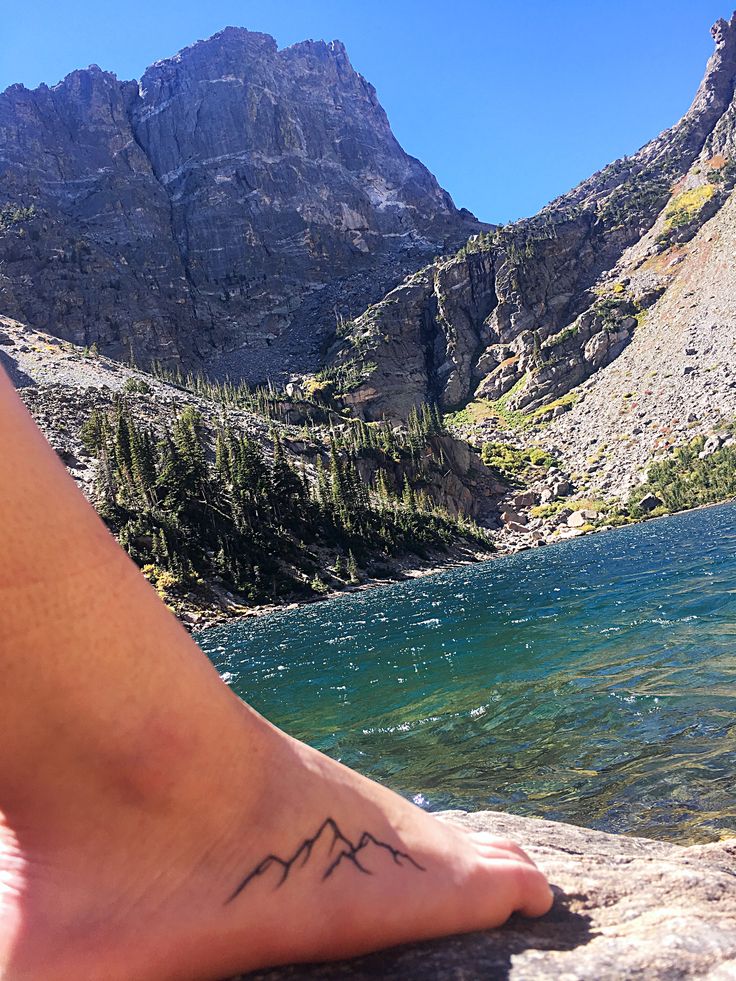 a person with a tattoo on their foot sitting next to the water in front of mountains