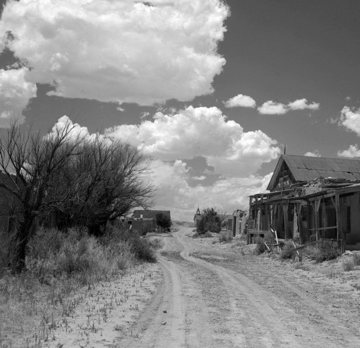 black and white photograph of an old house on dirt road with clouds in the sky