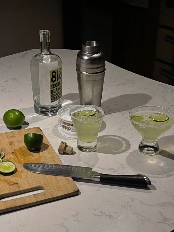 limes are sitting on the table next to two glasses and a cutting board with alcohol