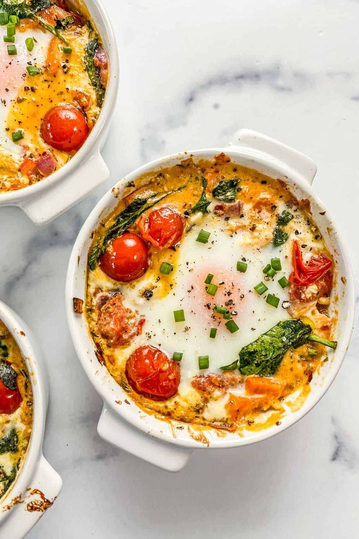 three white casserole dishes filled with eggs, tomatoes and spinach on a marble surface