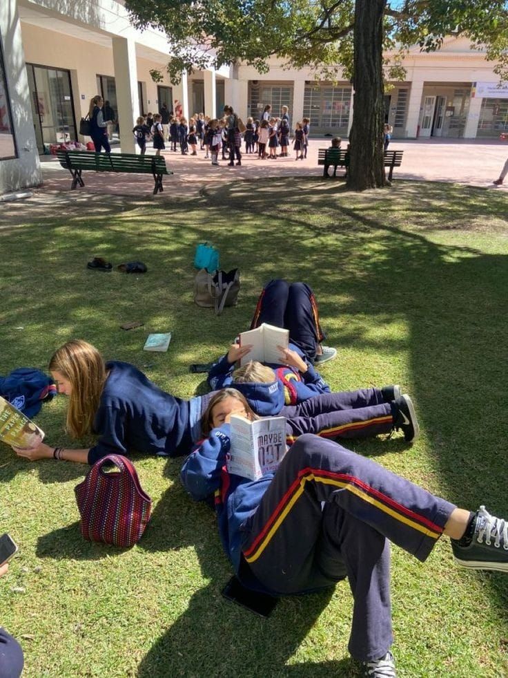 two people laying on the grass reading books in front of a building with students walking by