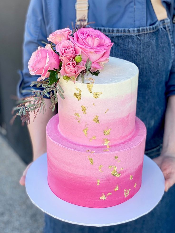 a person holding a pink and white cake with flowers on the top, while wearing overalls