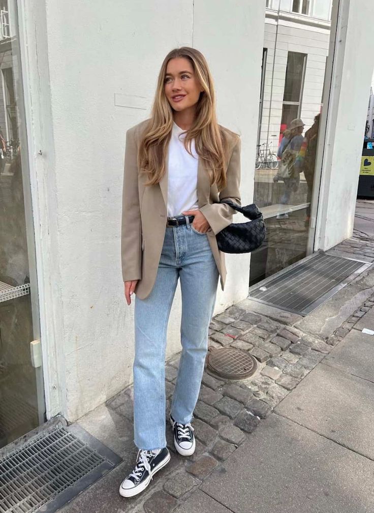 a woman standing in front of a building wearing jeans and a blazer with converse shoes