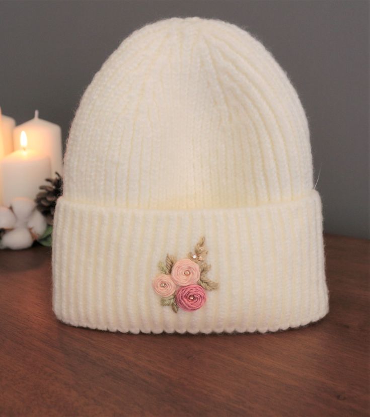 a white knitted hat with pink flowers on the front and side, next to two candles