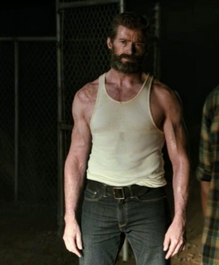 a man with a beard standing in front of a chain link fence wearing jeans and a tank top