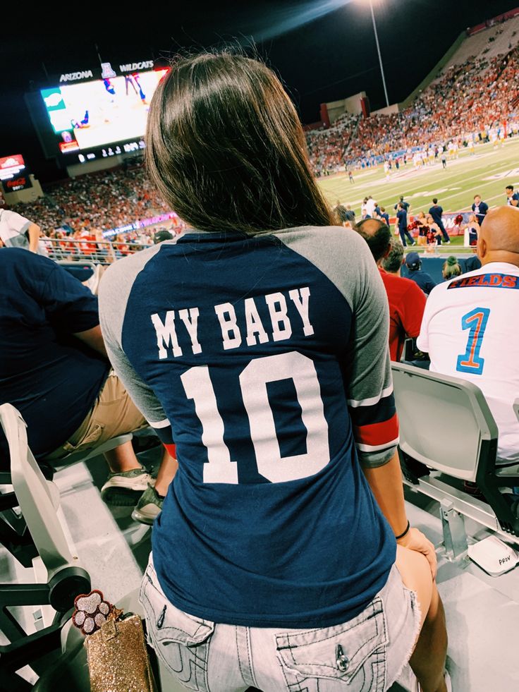 a woman sitting in the stands at a football game wearing a jersey that says my baby 10