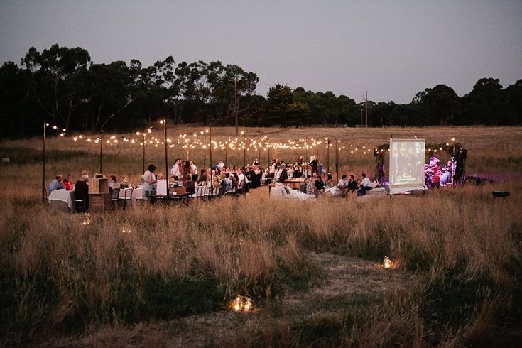 a large group of people sitting at tables in the middle of a field with lights strung over them