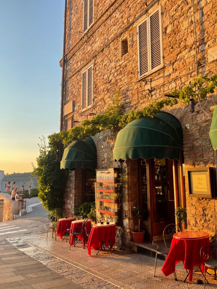 an old stone building with tables and chairs on the sidewalk in front of it at sunset