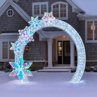 a lighted wreath in front of a house with snow on the ground and lights around it