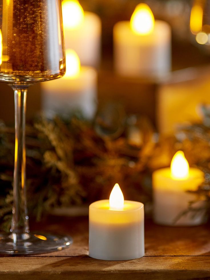 several lit candles on a table next to a wine glass and pine cones with evergreen branches in the background