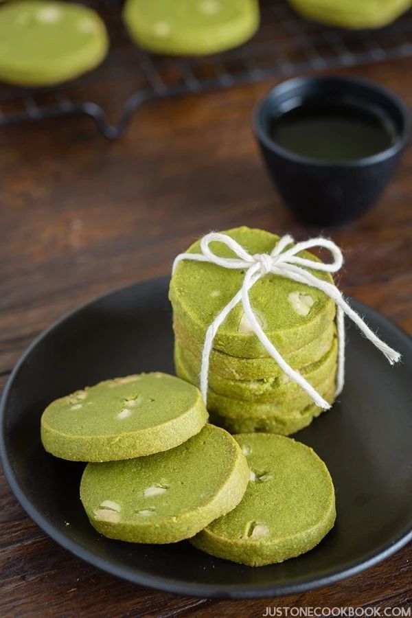 green tea cookies on a black plate with a white string tied around the top and two cups of coffee in the background