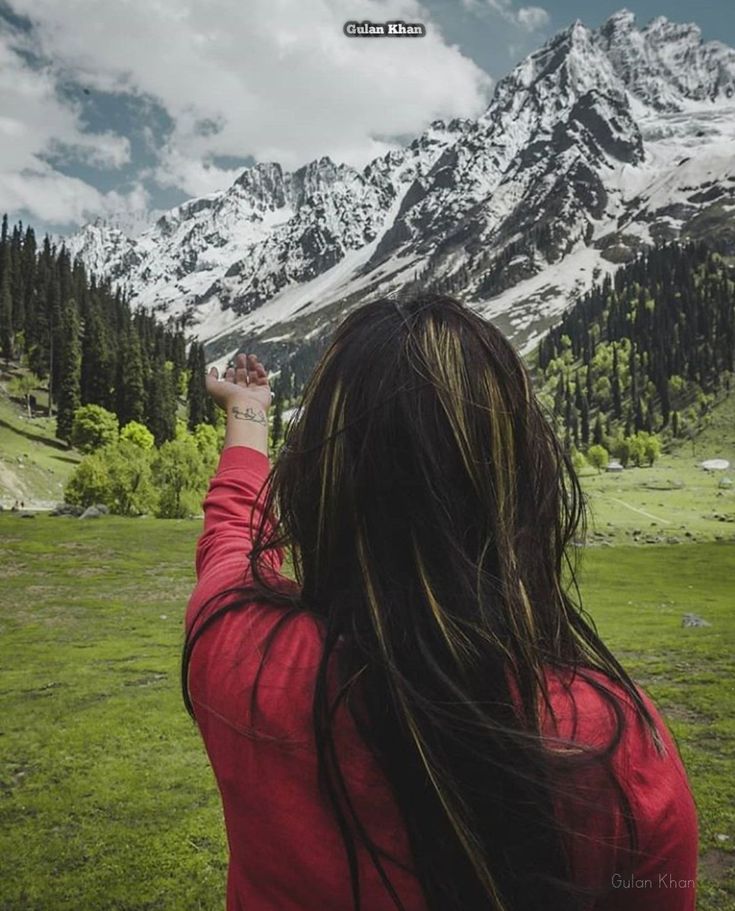 a woman in a red shirt flying a kite over a lush green field with snow covered mountains behind her