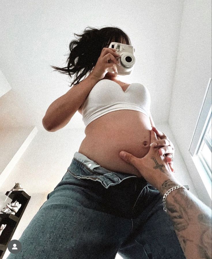 a pregnant woman taking a selfie with her camera in the mirror while wearing jeans