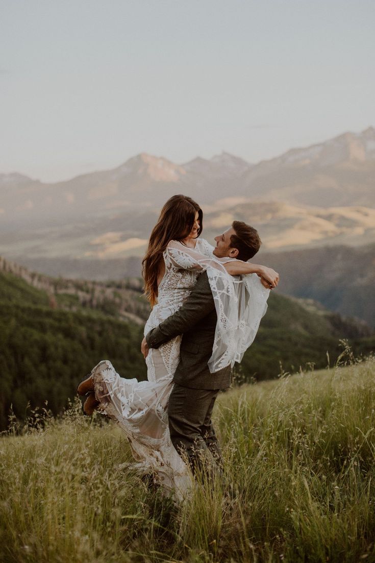 a man carrying a woman in his arms while standing on top of a grass covered hill