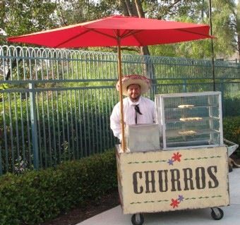 a man with a hat and tie standing behind a cart that says churros