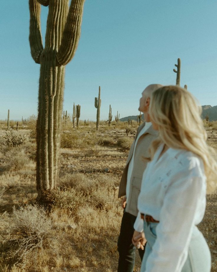 a man and woman standing in front of a large saguado cactus on a sunny day