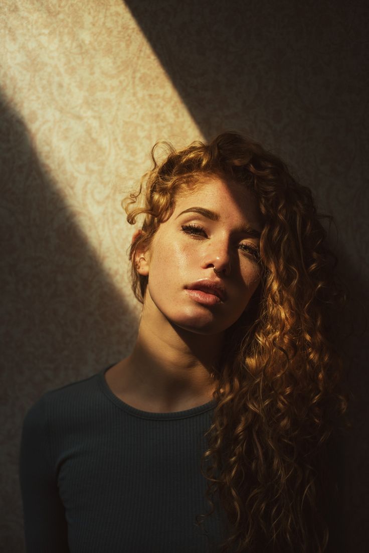 a woman with curly hair standing in front of a wall and looking at the camera