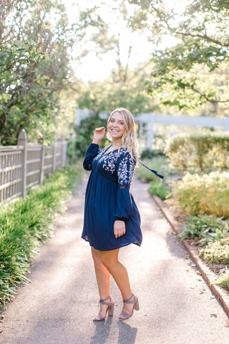 a woman in a blue dress poses for a photo on a path with trees and bushes behind her