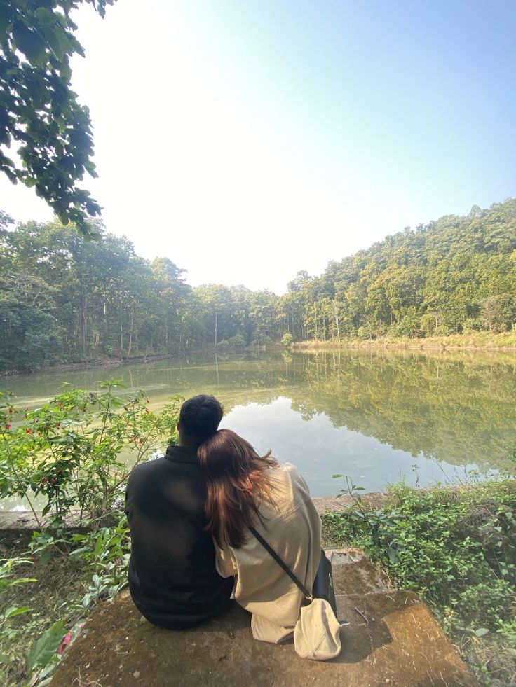 a woman sitting on top of a rock next to a lake with trees in the background