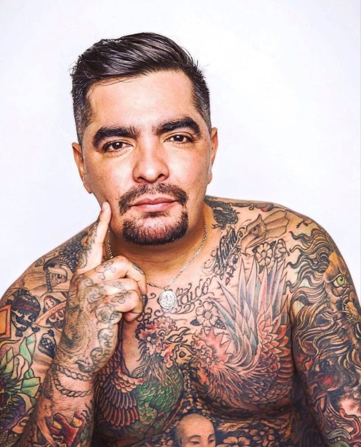 a man with lots of tattoos on his chest posing for the camera while holding his finger to his chin