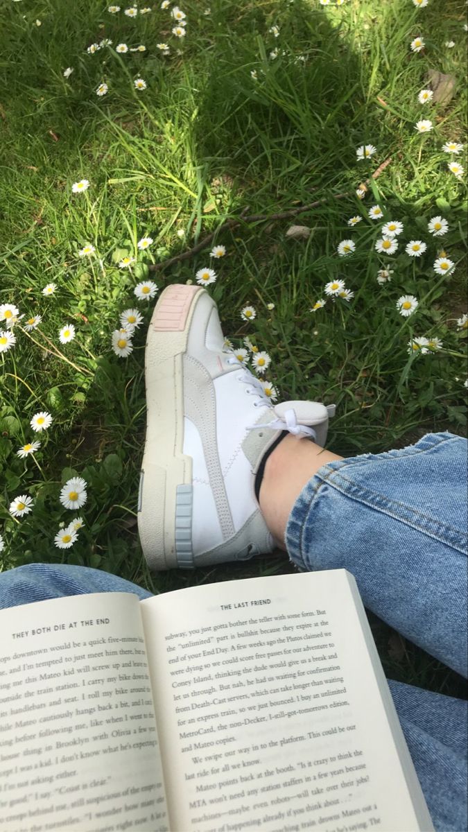 a person sitting in the grass with their feet crossed and reading a book next to daisies