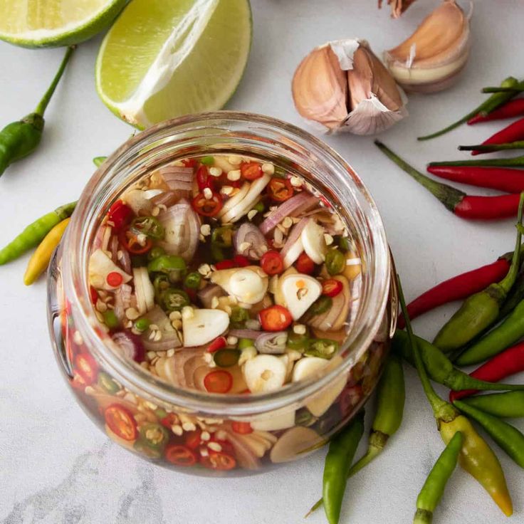 a glass jar filled with vegetables next to green peppers and lime wedges on a white surface