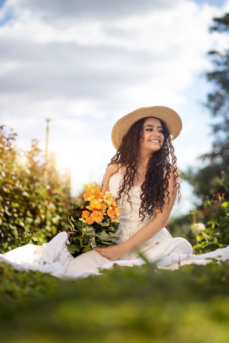 a woman sitting on the ground with flowers in her hand and wearing a straw hat