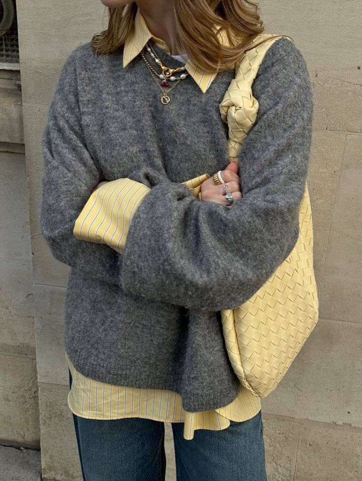 a woman standing in front of a building with her hand on her hip, wearing a gray sweater and blue jeans