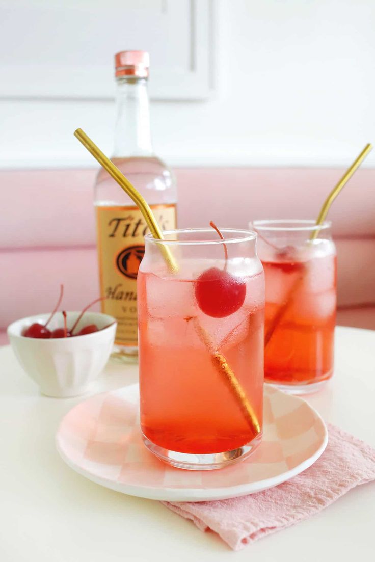 two glasses filled with pink lemonade and cherries on a plate next to a bottle of booze