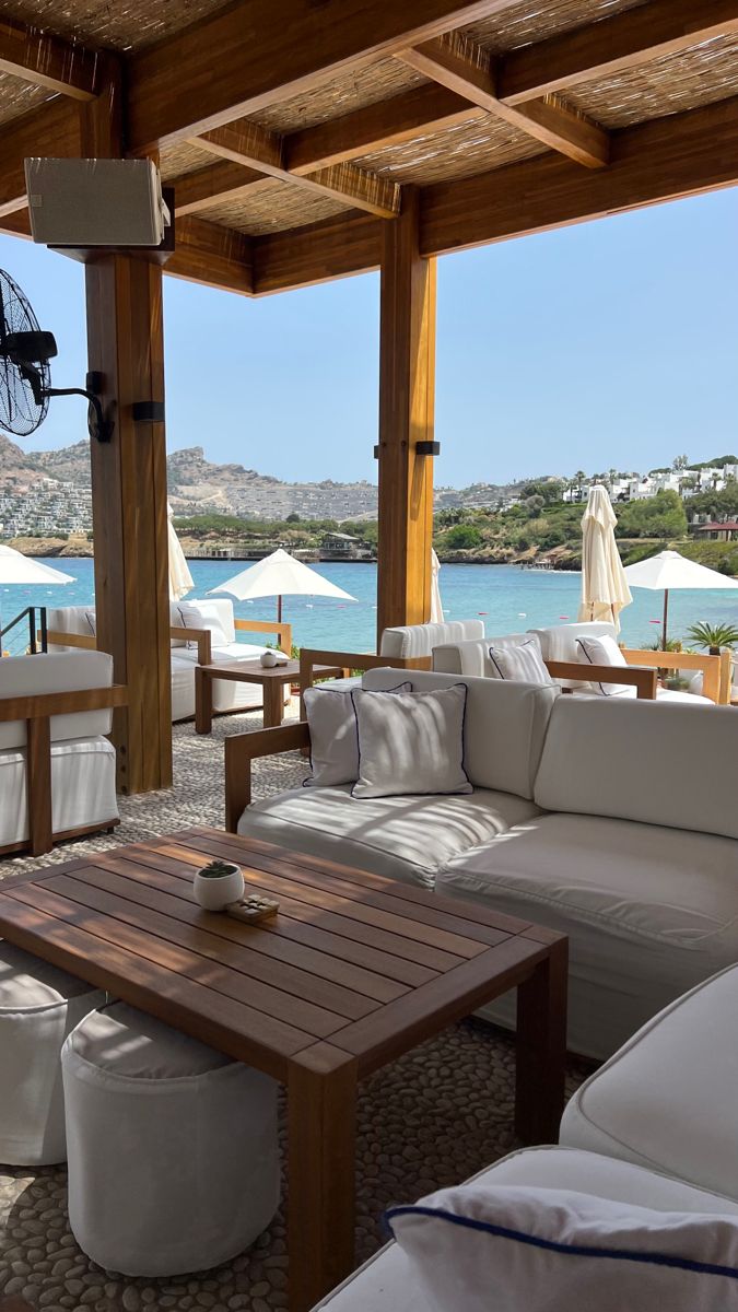 an outdoor living area with white furniture and umbrellas over looking the water, on a sunny day