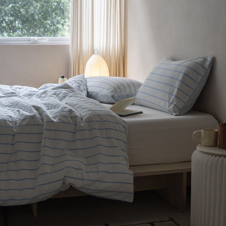 an unmade bed with blue and white striped comforter next to a window in a bedroom