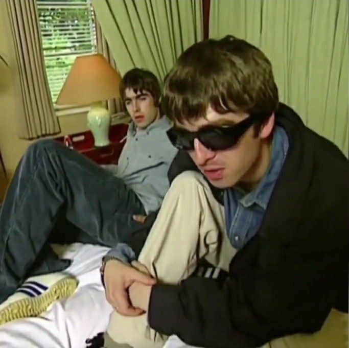 two young men sitting on a bed with one looking at his cell phone while the other looks down