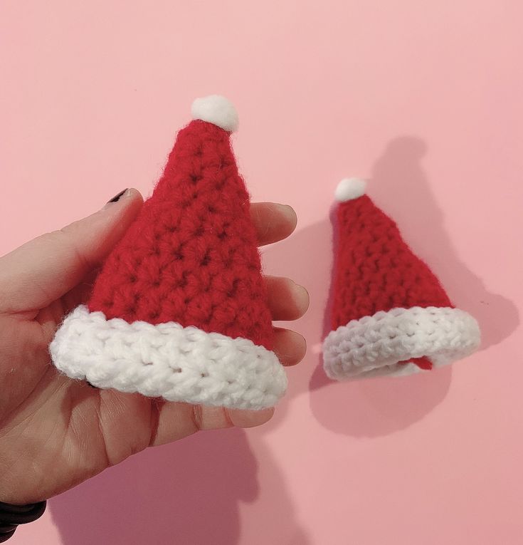 a hand holding two crocheted santa hats on top of each other in front of a pink background