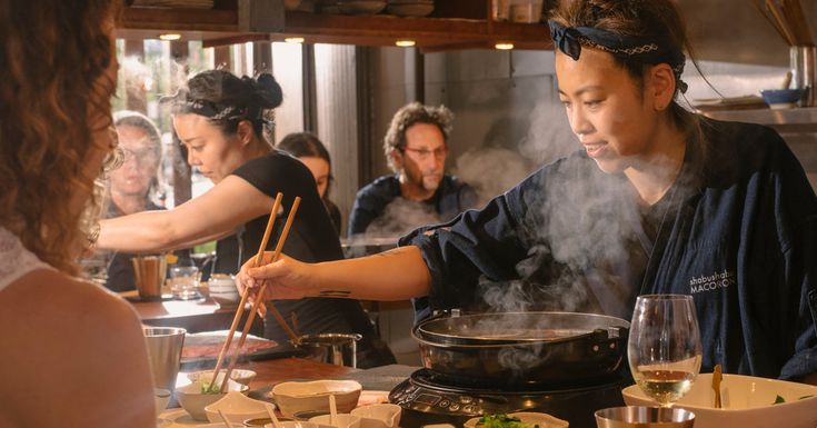 a group of people standing around a table with food on it and chopsticks in their hands