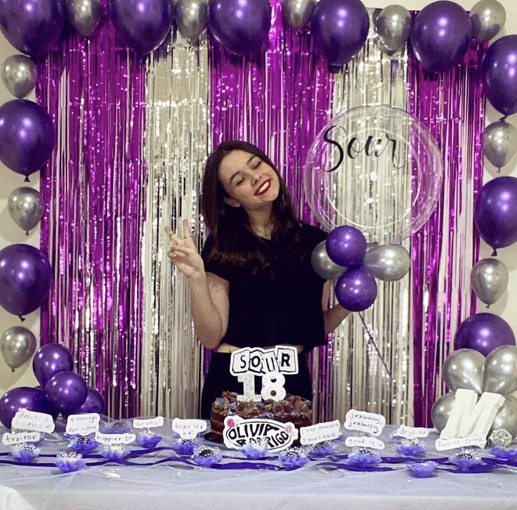 a woman standing in front of a table with purple and silver balloons, cake and streamers