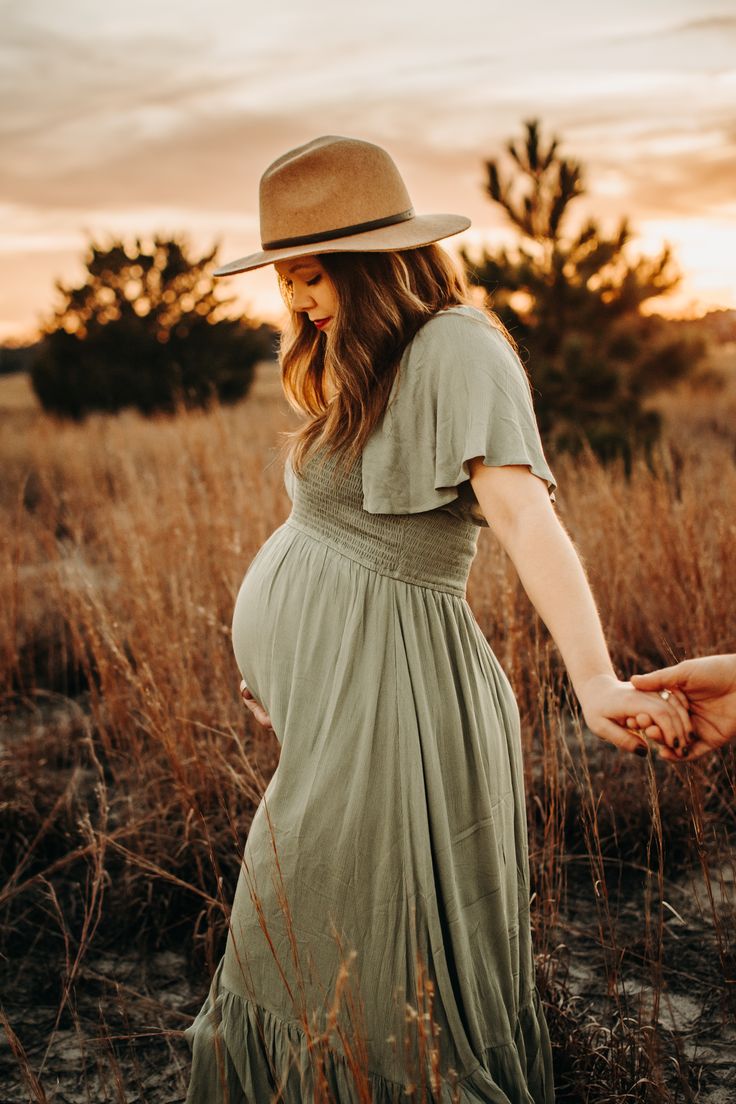 a pregnant woman in a dress and hat walks through tall grass with her hands behind her back